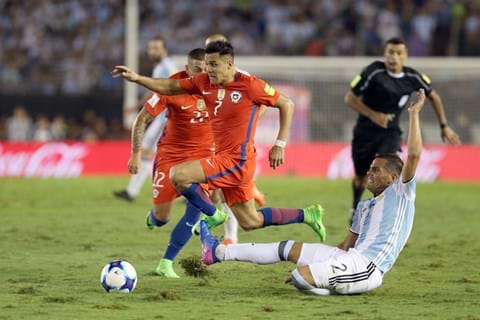 Chilean footballer running past an Argentina player attempting a slide tackle, with another player and referee in the background.